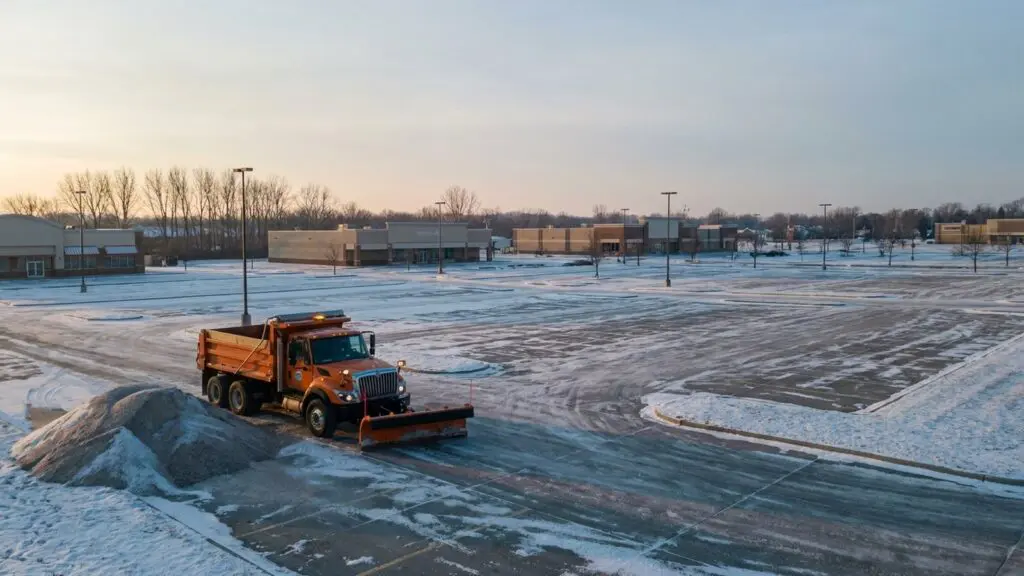Snowplow truck idling beside a salt pile at a snow-dusted Omaha commercial plaza at sunrise